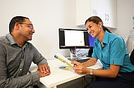 A female clincian leaning over her desk holding a booklet and talking to a male patient